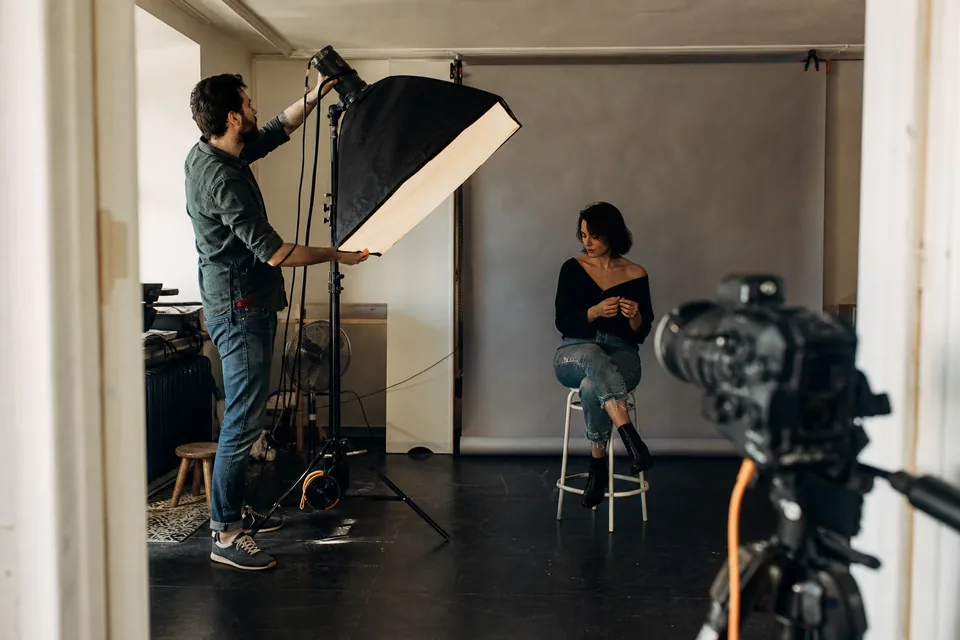Fotograf ustawia światło w studiu przed sesją portretową, modelka czeka na stołku